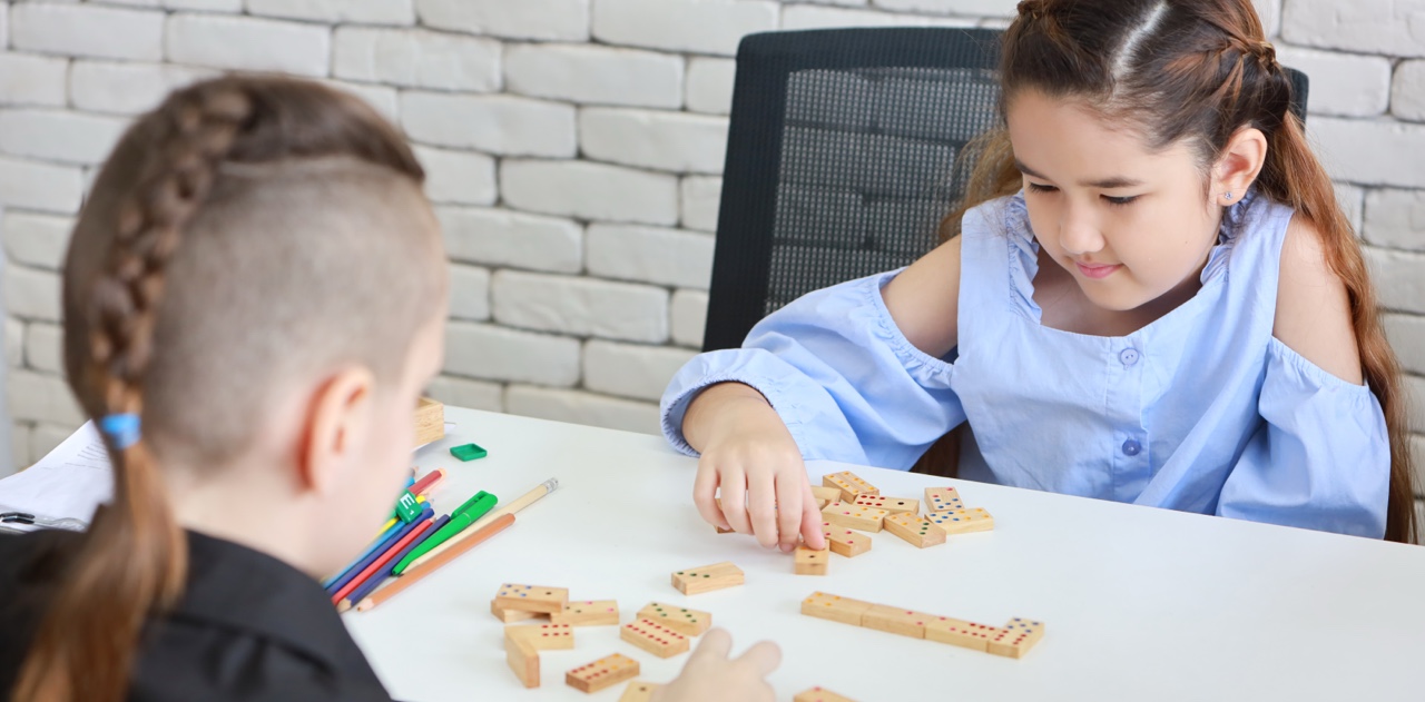 Two children playing a game of wooden dominoes at a classroom table, practicing strategy and learning through play.